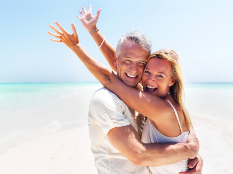 Smiling middle-aged couple embracing on a sunny beach, looking happy and carefree, with clear blue water and sky in the background.