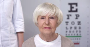 A senior woman stares into the camera. An eye exam chart is out of focus behind her.