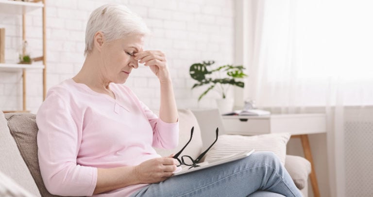 A senior woman holds her glasses on her lap and pinches the bridge of her nose in discomfort