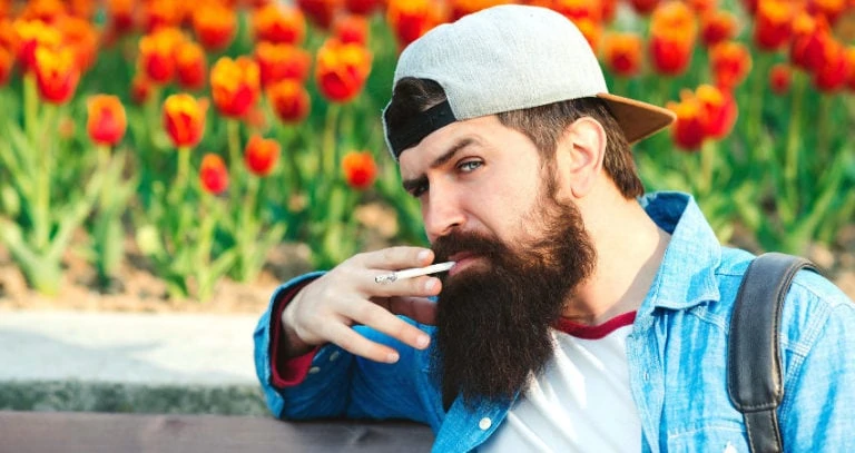 A man with a beard squints at the camera and smokes a cigarette