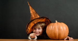 A child wearing a Halloween witch hat sits next to a pumpkin and points at the camera.