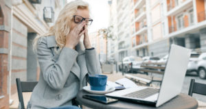 A woman pushes her glasses off the bridge of her nose and rubs her eyes. She is seated at an outdoor cafe table working on her laptop