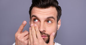 A man holds his cheek with one hand and holds a contact lens in the other hand.