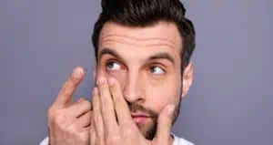 A man holds his cheek with one hand and holds a contact lens in the other hand.