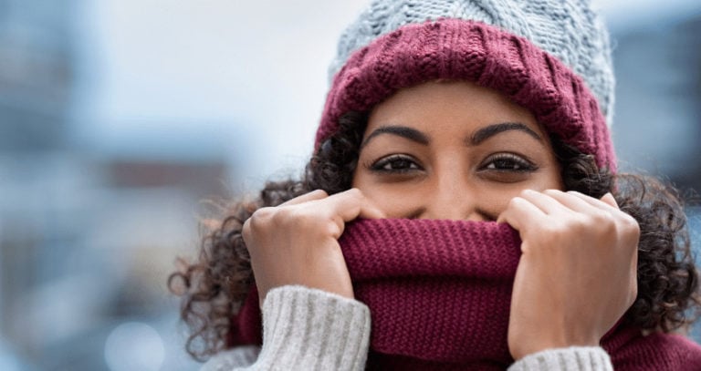 A woman with a knit hat on holds a scarf above her nose and grins.