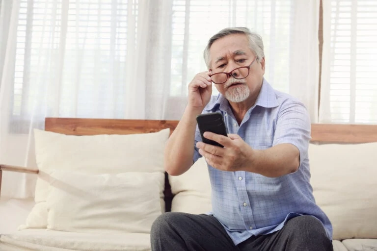 An older man holds his glasses against his face and looks at his phone screen for cataract surgery information