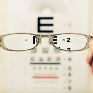 Someone holds a pair of glasses in front of an eye exam chart. The area outside of the glasses is blurry, but the area inside the lenses is clear