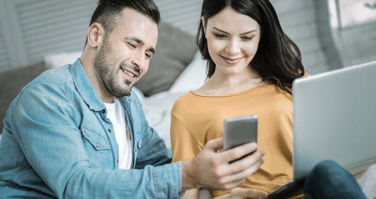 A young man and woman smile at a cell phone. The woman is holding a laptop as well.