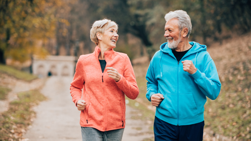 An older couple jogging together on a park path in autumn, smiling at each other while wearing colorful athletic jackets.