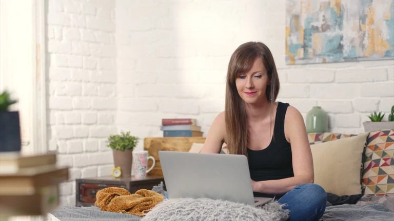 A woman sits on her bed and smiles at her laptop