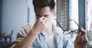 A young man is holding his glasses away from his face as he pinches the bridge of his nose in discomfort