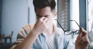 A young man is holding his glasses away from his face as he pinches the bridge of his nose in discomfort