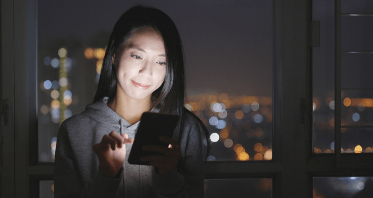 A young woman in a dark apartment looks at her bright phone screen