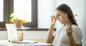 A young woman holds her glasses and rubs her eye as she looks at her laptop.