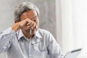 An elderly man holds his glasses and rubs his eyes while trying to read something