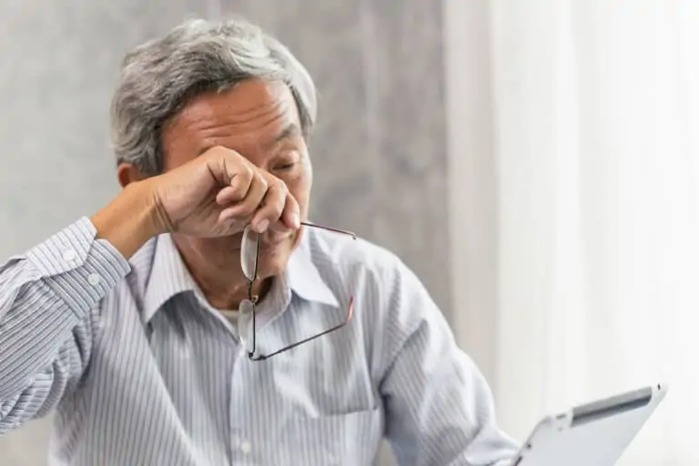 An elderly man holds his glasses and rubs his eyes while trying to read something