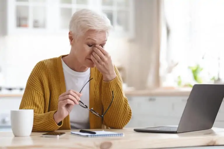 A senior woman holds her glasses and pinches the bridge of her nose in discomfort