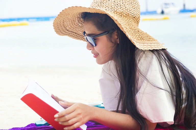 A young woman wearing sunglasses reads a book on the beach