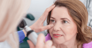 An older woman stares into an eye-examining device. The eye exam is being performed by a blonde eye doctor