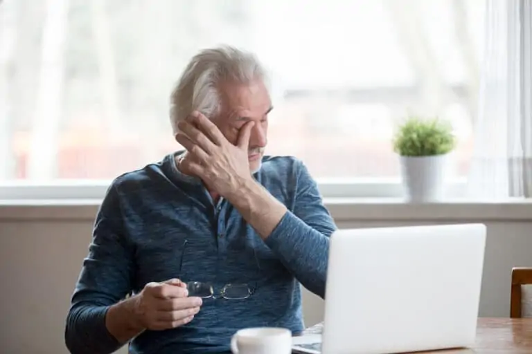 A man holds his glasses and rubs his eyes in discomfort as he uses a computer