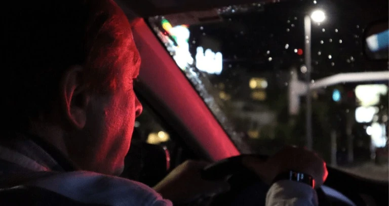 Older man drives his car at night, a red stoplight reflects on his face.