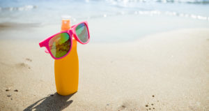 A sunscreen bottle is sitting in the sand at a beach. A pair of sunglasses has been placed on top of the bottle