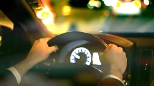 A man's hands grip a car's steering wheel while he faces a blurry road at night.