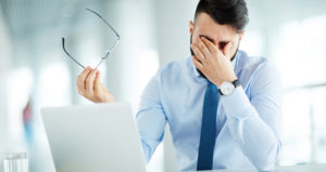 A man holds his glasses away from his face and rubs his eyes. He is sitting with a laptop