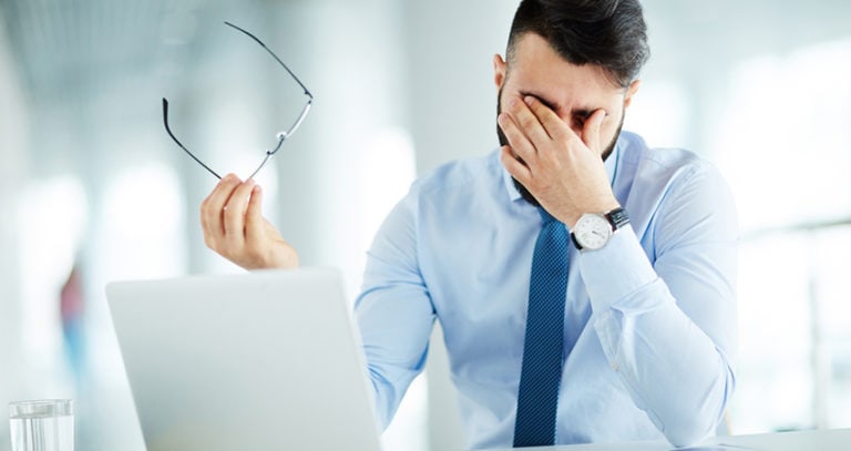 A man holds his glasses away from his face and rubs his eyes. He is sitting with a laptop