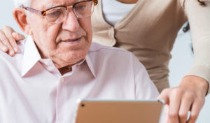 An older man with glasses looks at an iPad. A younger woman behind him points to something on the iPad.