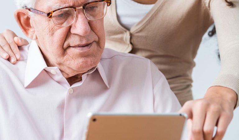 An older man with glasses looks at an iPad. A younger woman behind him points to something on the iPad.