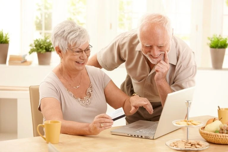 A smiling older couple is looking at a laptop screen. The woman wears glasses and is holding a credit/debit card.