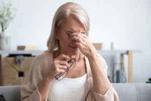 An older woman is pinching her eyes in discomfort. She is holding her glasses in one hand