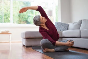 An active, older woman performs stretching exercises on a yoga mat in her living room.